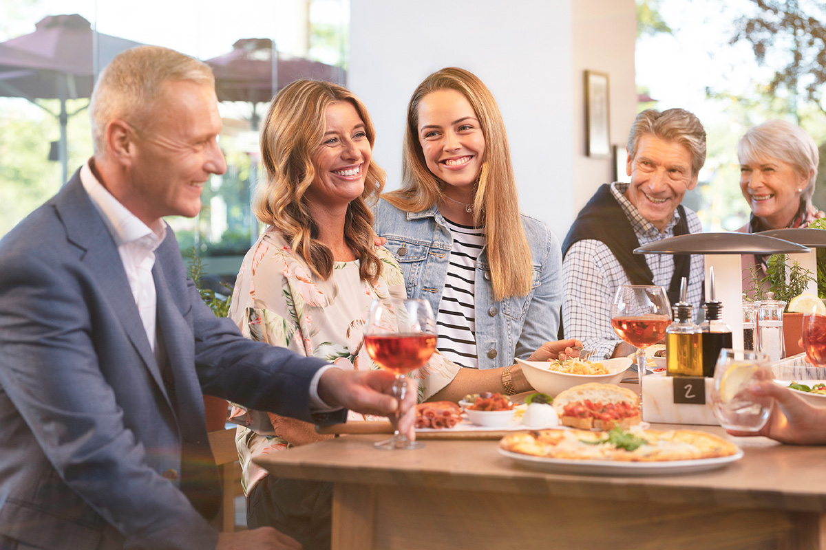 Five people smiling and enjoying a meal with wine at a restaurant table.
