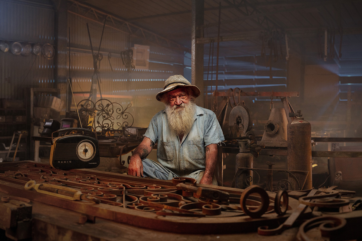 Bearded elderly man wearing a hat and blue shirt sitting in a workshop with metal tools and vintage radio.