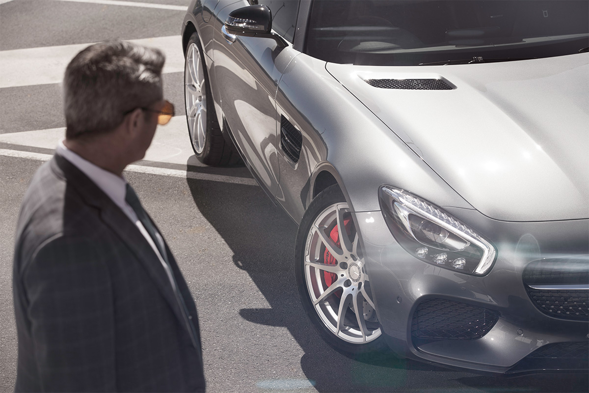 Man in suit and sunglasses looking at a gray sports car parked on asphalt.
