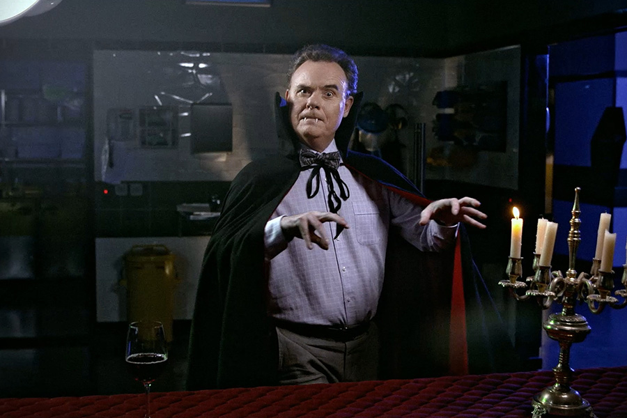 Man in vampire cape with bow tie, fangs, and claws, standing beside lit candelabra and wine glass.