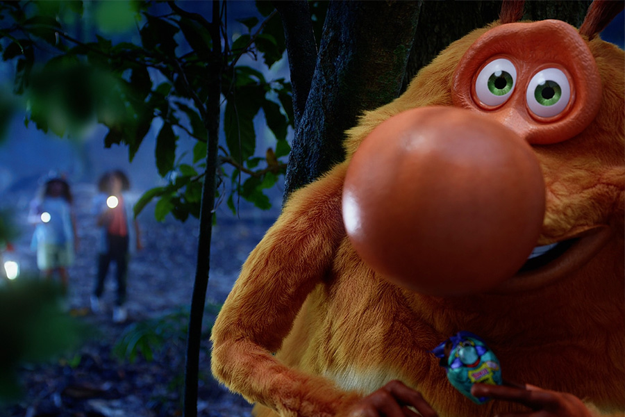 Large orange cartoon creature with green eyes holding candy hiding behind a tree at night.