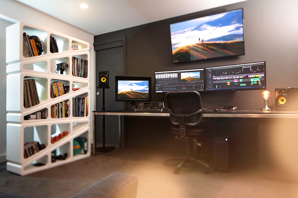 Modern editing workspace with three monitors, speakers, a chair, and a unique white bookshelf filled with items.
