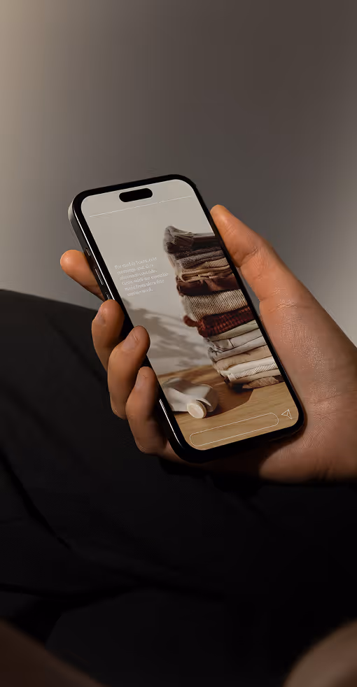 A hand holding a smartphone displaying the Timininous mobile interface. The screen shows folded clothing stacked on a wooden surface with soft lighting and minimal interface elements.
Mockup.Maison