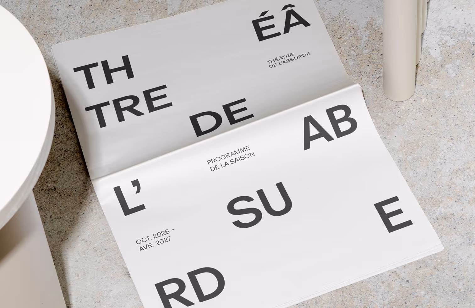 A folded theatre programme placed on a textured surface featuring large fragmented typography across the cover. The layout combines bold letterforms with minimal text to present the Théâtre de l’absurde seasonal programme.

Mockup from Bendito Mockup
