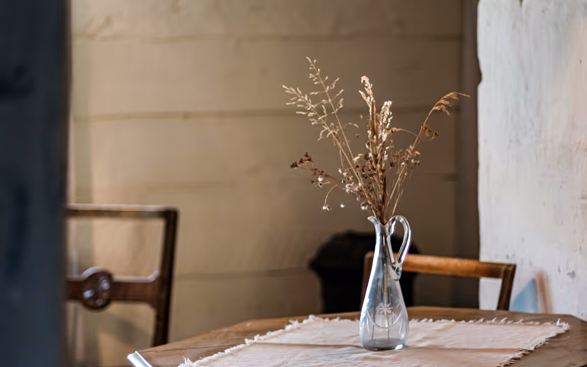 A softly lit interior scene featuring dried flowers arranged in a glass vase on a wooden table. The warm tones, natural textures, and quiet composition create a calm and intimate domestic atmosphere.