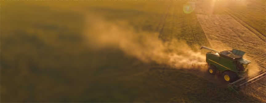 A green combine harvester moves through a golden field kicking up dust at sunset.