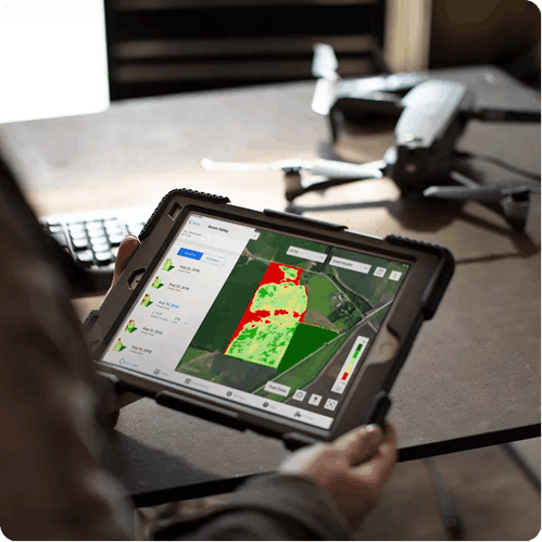 Person holding tablet showing color-coded agricultural field map with drone on table nearby.