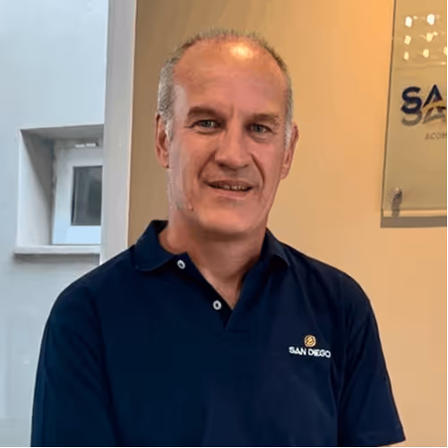 Man in dark blue San Diego polo shirt smiling indoors near a window and wall sign.