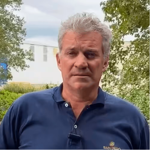 Man with gray hair wearing a navy blue San Diego polo shirt outdoors near trees and a building.