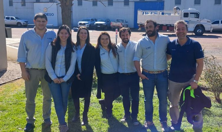 Siete personas posan juntas al aire libre frente a un edificio industrial y árboles sin hojas.