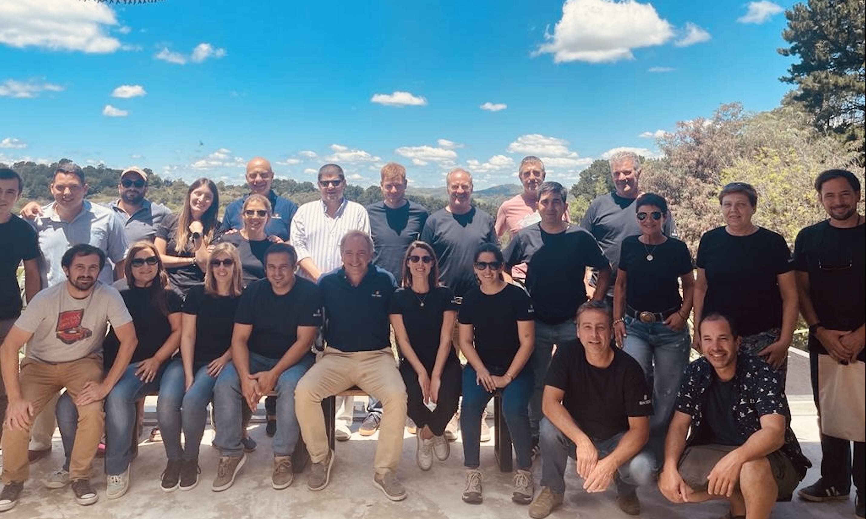 Grupo de 24 personas posando al aire libre con cielo azul y árboles al fondo.