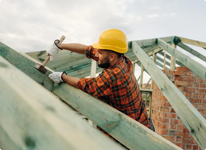 Construction worker wearing a hard hat working on wooden roof framing at a job site.