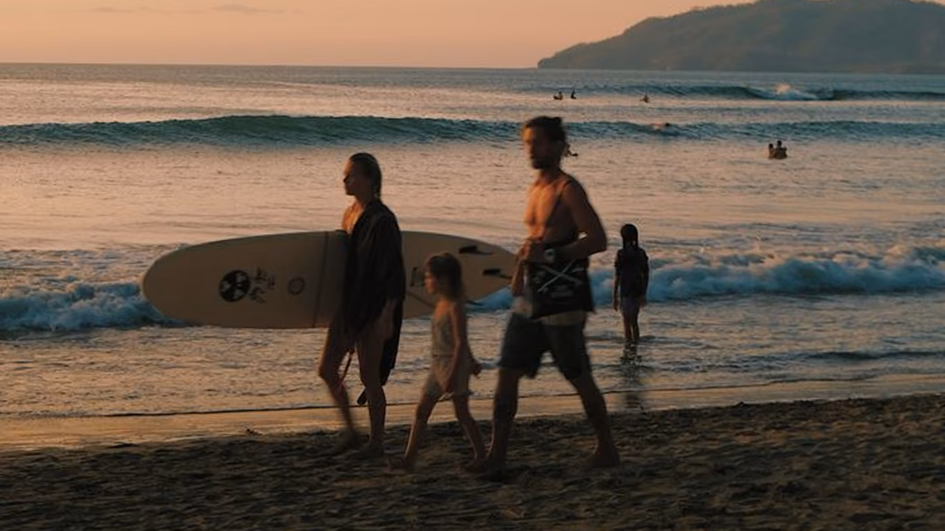 Family walking on the beach at sunset with a surfboard and ocean waves in the background.