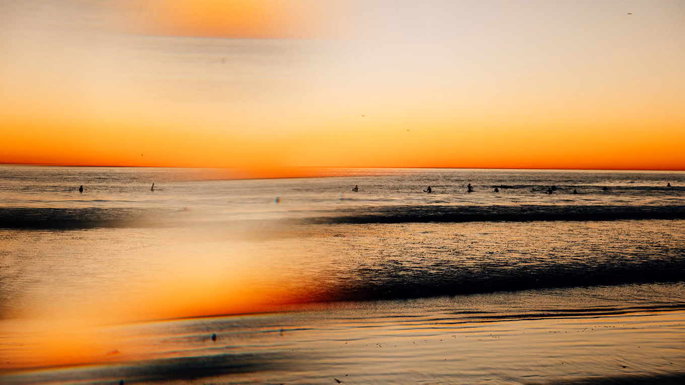 Surfers in the ocean during a vibrant orange sunset with reflections on the water and beach.