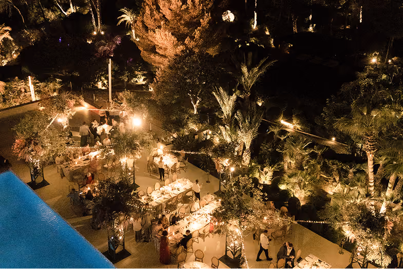 Evening outdoor dining event with guests at lit tables surrounded by illuminated trees and plants.