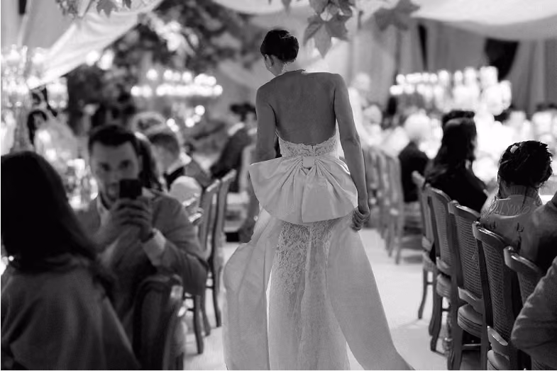 Bride in lace gown with large bow walks down aisle past seated guests at an indoor wedding reception.