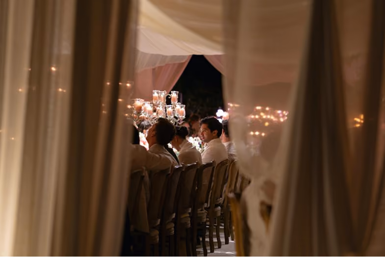 People seated at a long dining table with lit candles, viewed through translucent curtains.