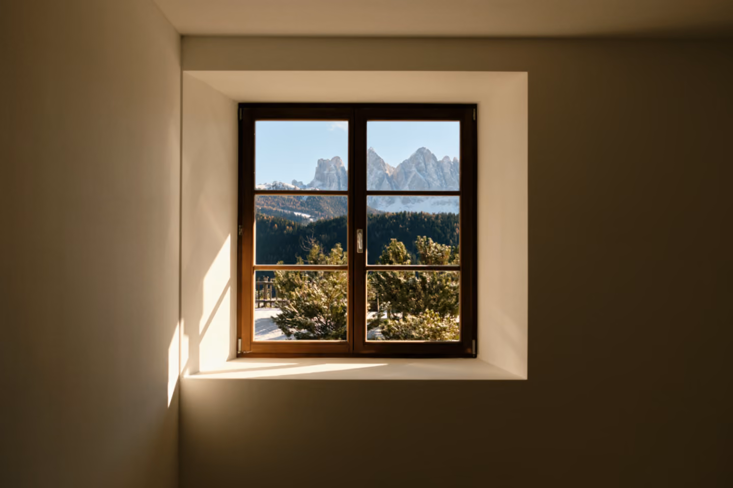 View through window showing pine trees and snow-capped mountains under blue sky.