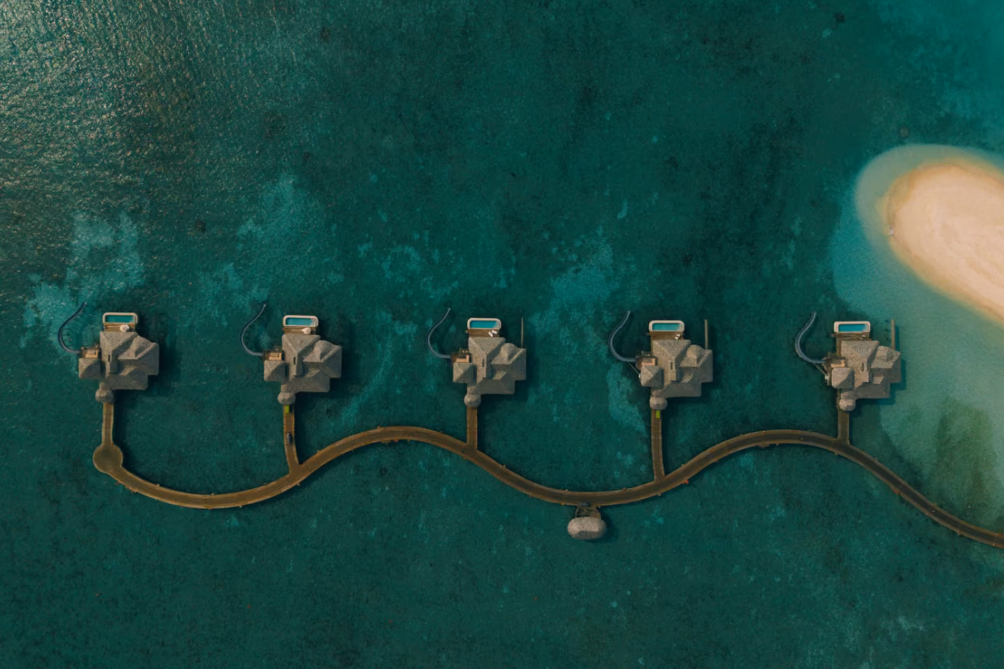 Aerial view of five overwater bungalows connected by a curved wooden walkway over turquoise ocean near a sandy shore.
