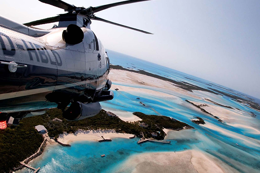 View of a helicopter flying over turquoise waters and sandy islands with docks and greenery below.