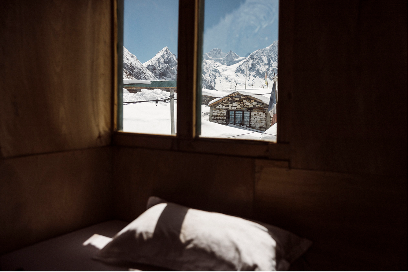 View of snowy mountains and a stone building through a window above a bed with a pillow.