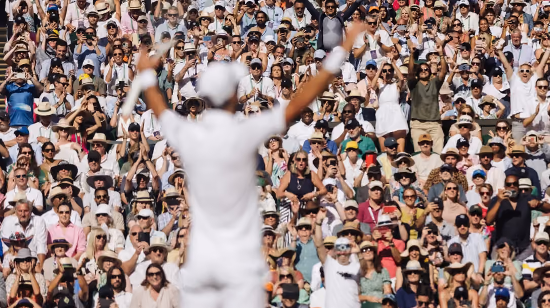A cricket player in white uniform with arms raised facing a large crowd of diverse spectators, many of whom are clapping and cheering in an outdoor stadium.