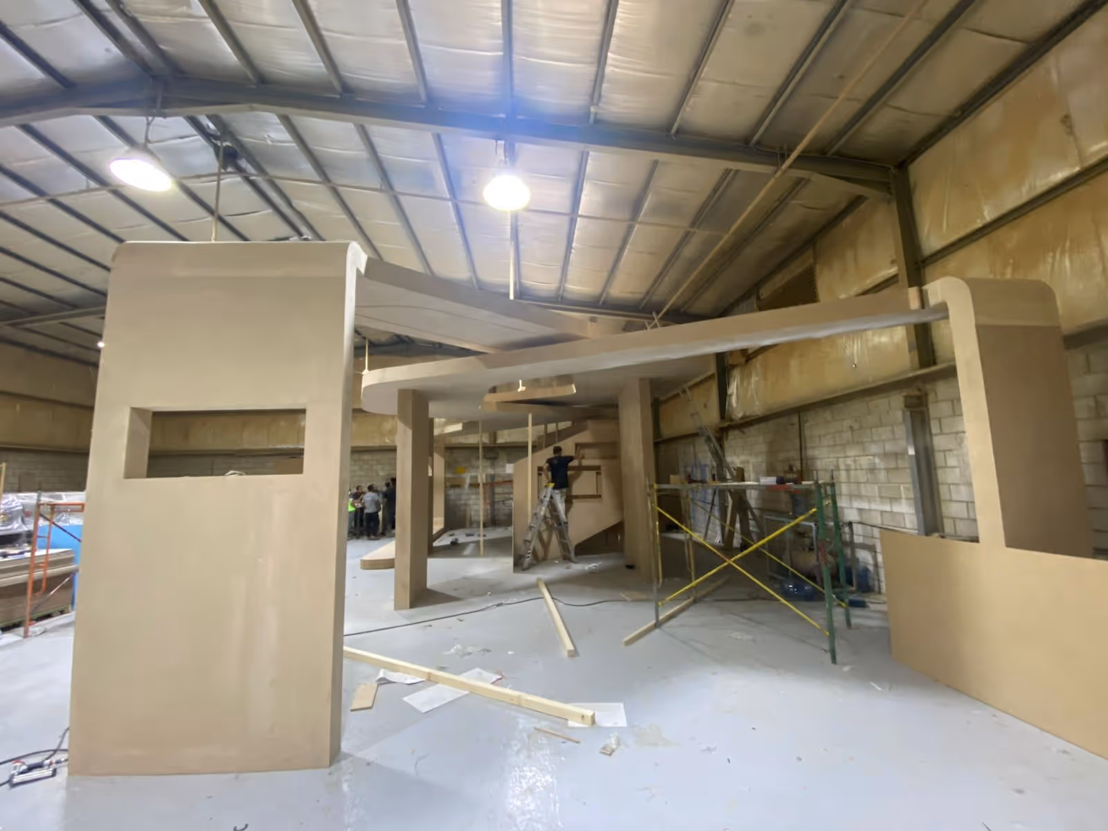 Large unfinished wooden structure inside warehouse with workers and construction materials around.