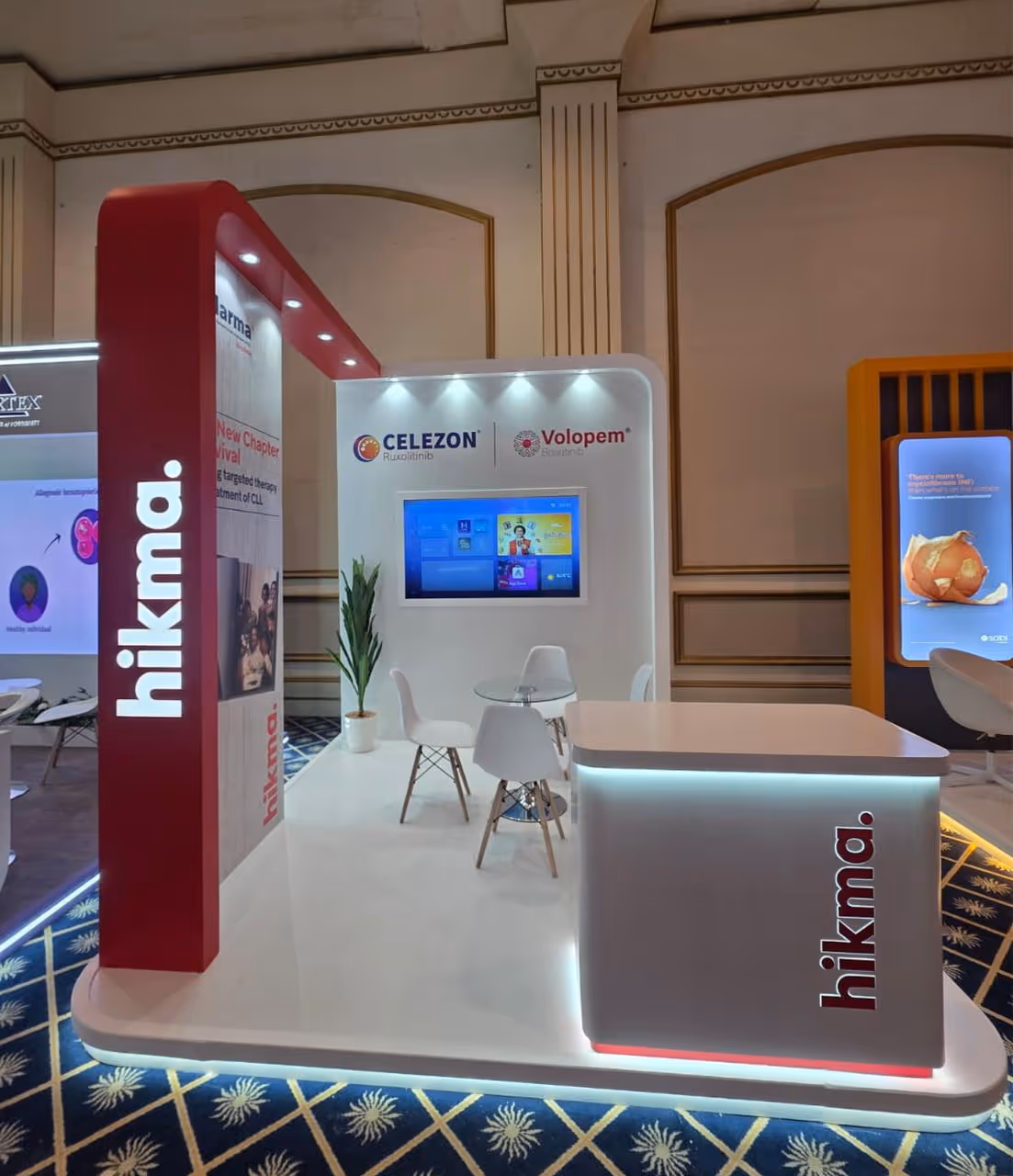 Hikma pharmaceutical booth with illuminated signs, chairs, table, plant, and a monitor on a decorated carpet floor.