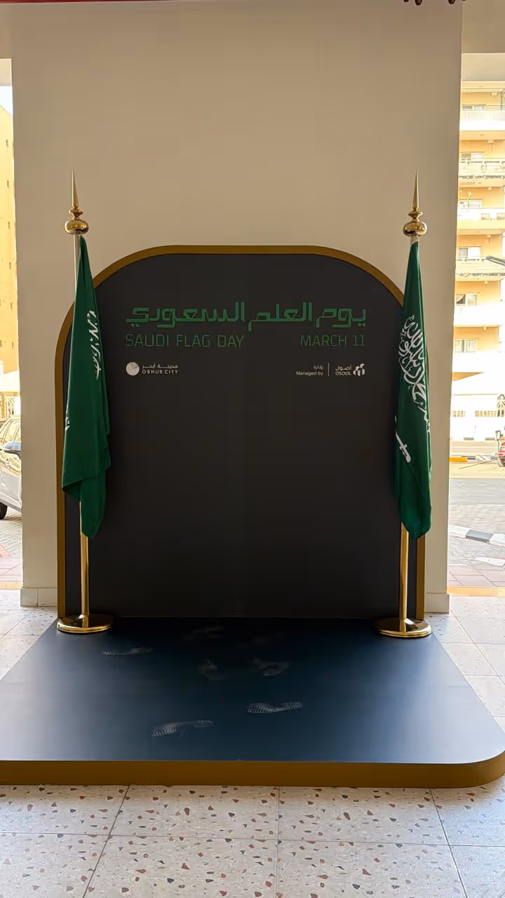 Two Saudi flags on stands flanking a black display for Saudi Flag Day, March 11, with logos below.