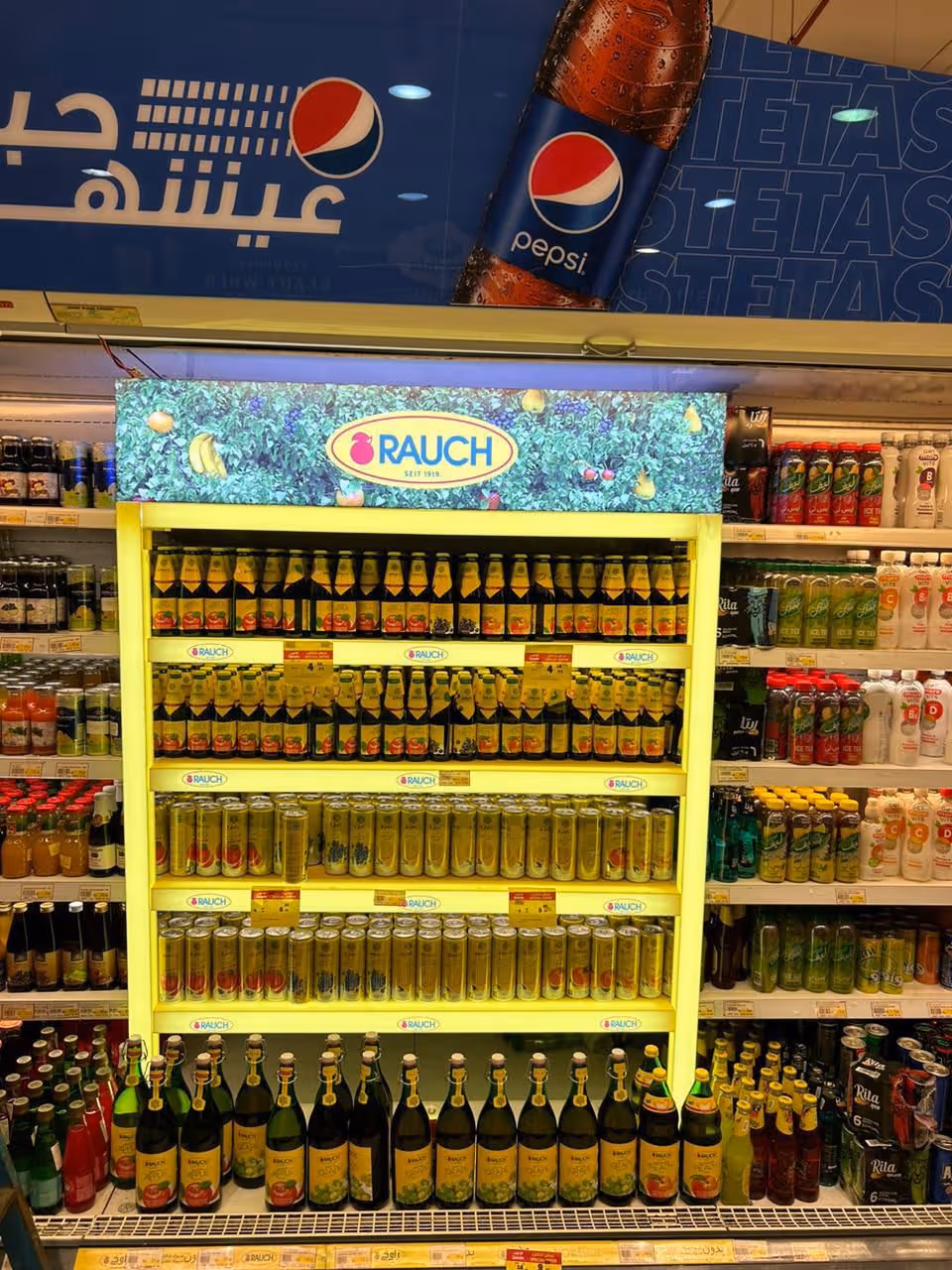 Supermarket shelf stocked with various Rauch brand juice bottles and cans lit by yellow shelf lights.