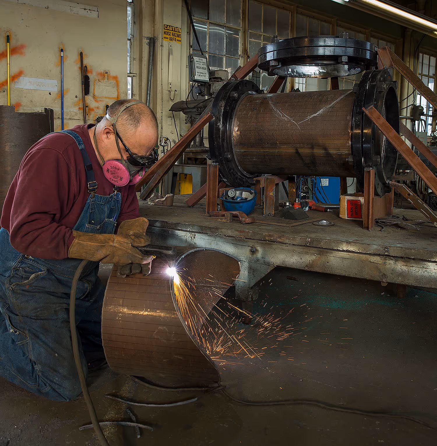 Worker wearing goggles and gloves cutting metal pipe with a torch producing sparks.