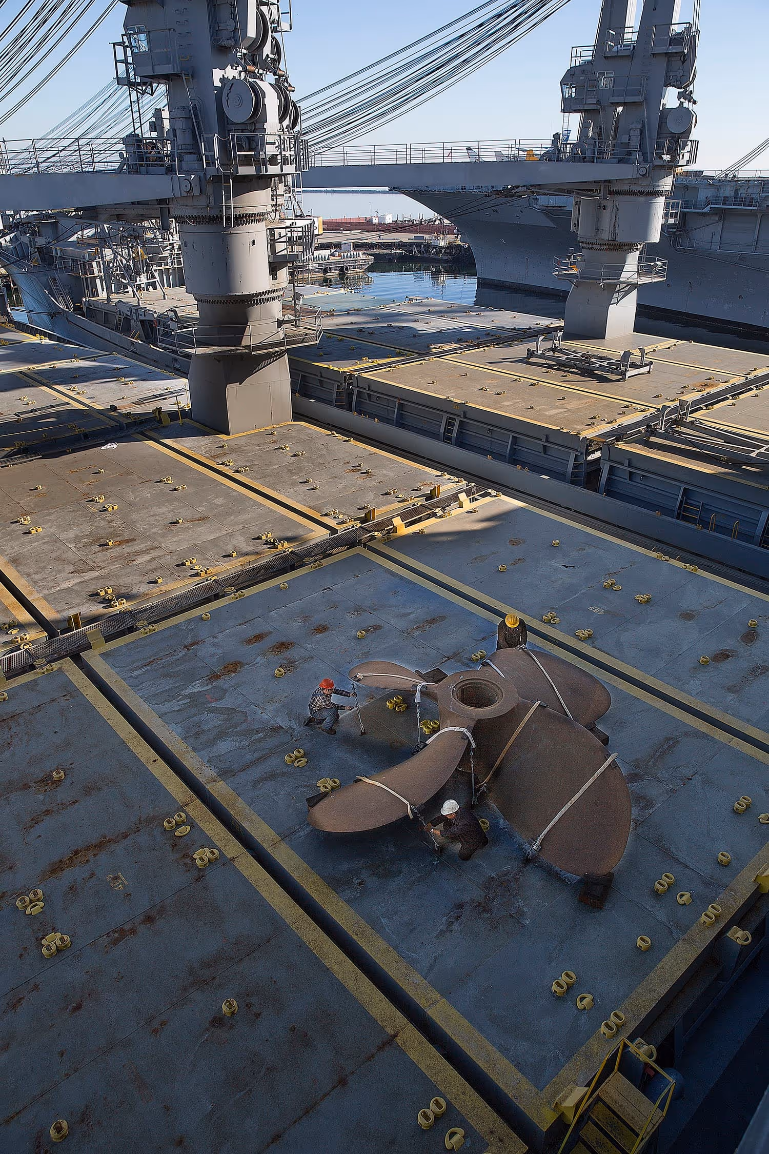 Three workers in hard hats securing a large metal ship propeller on a ship's deck with cranes overhead.
