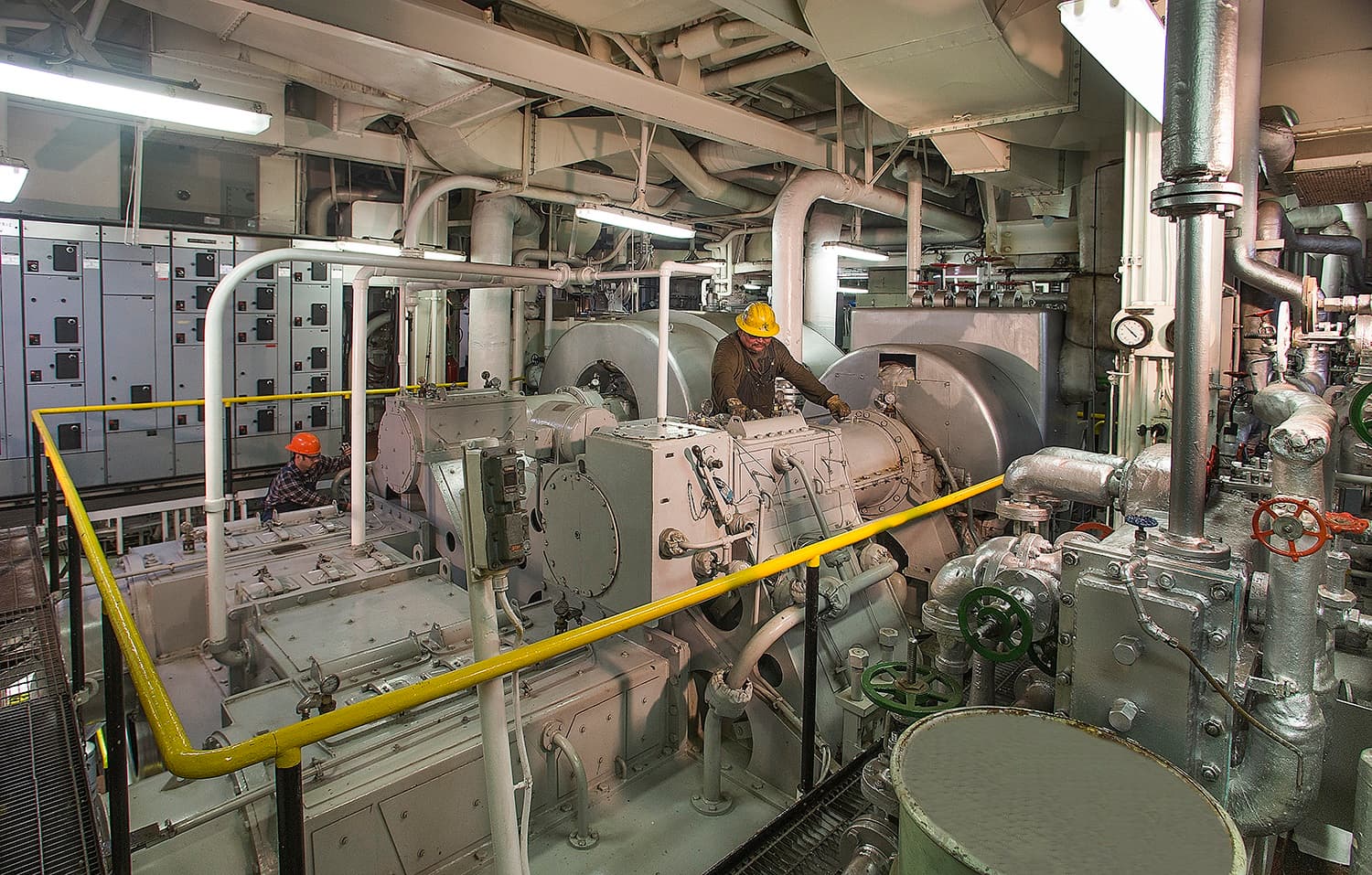 Two workers wearing helmets operating large industrial machinery in a factory plant room.