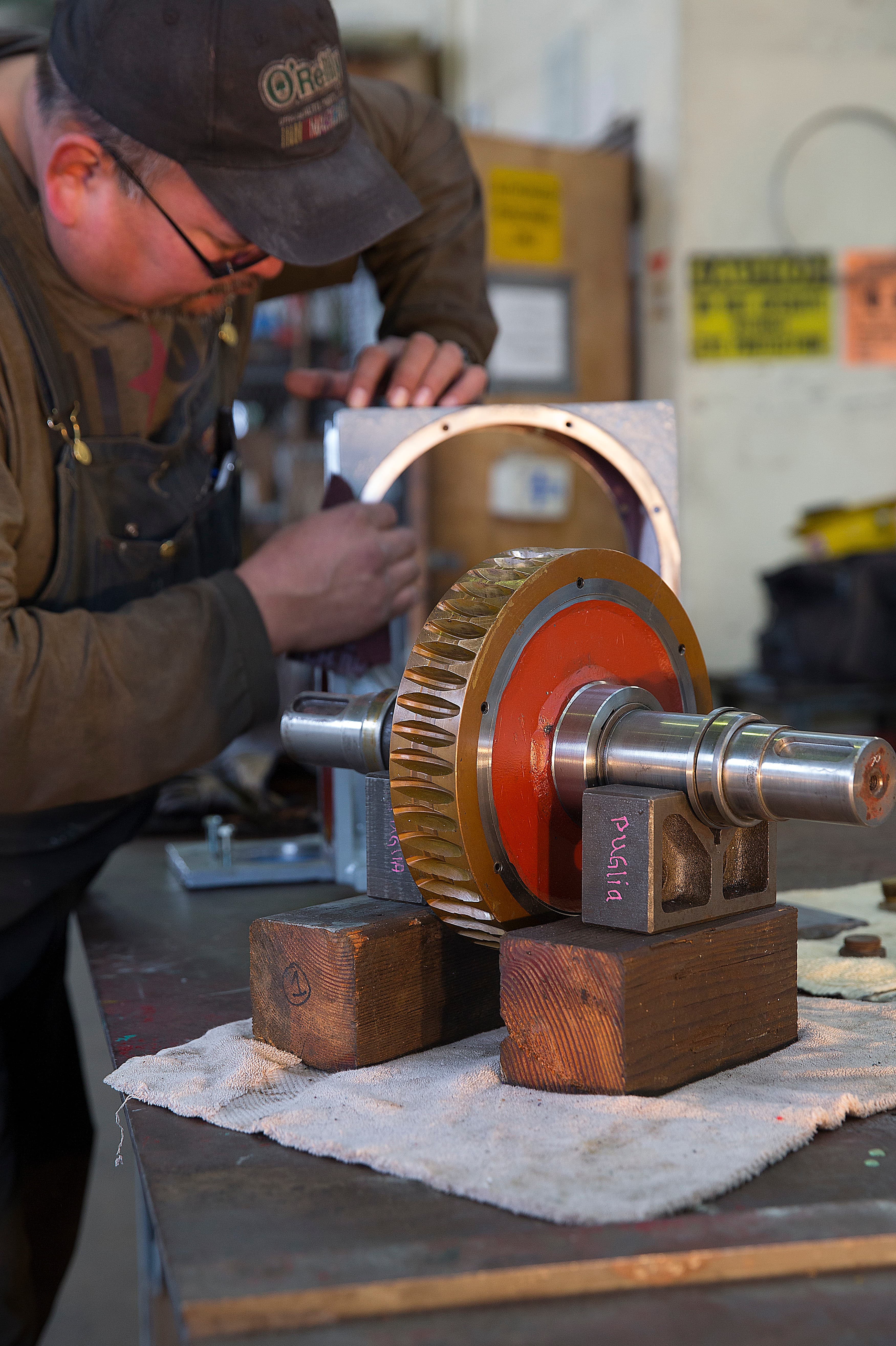 Man in cap working on a metal gear assembly placed on wooden blocks in a workshop.