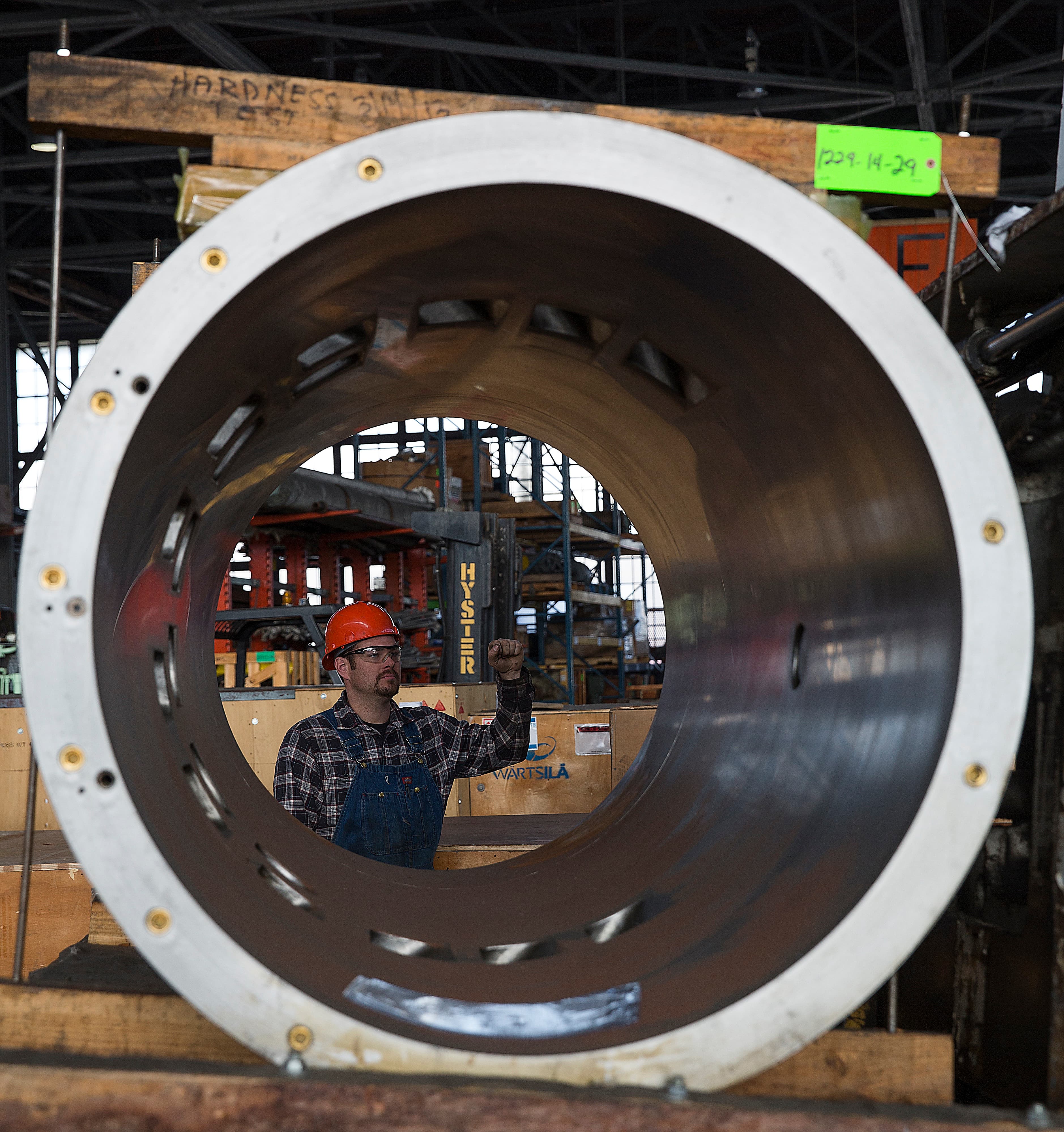 Worker in protective gear pictured through the opening of a large metal pipe in a factory.