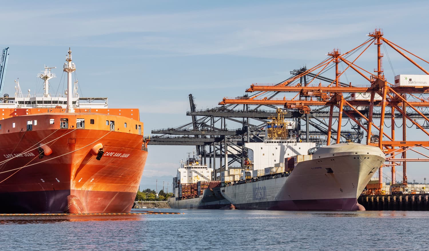 Large orange and white cargo ships docked at a busy port with industrial cranes.