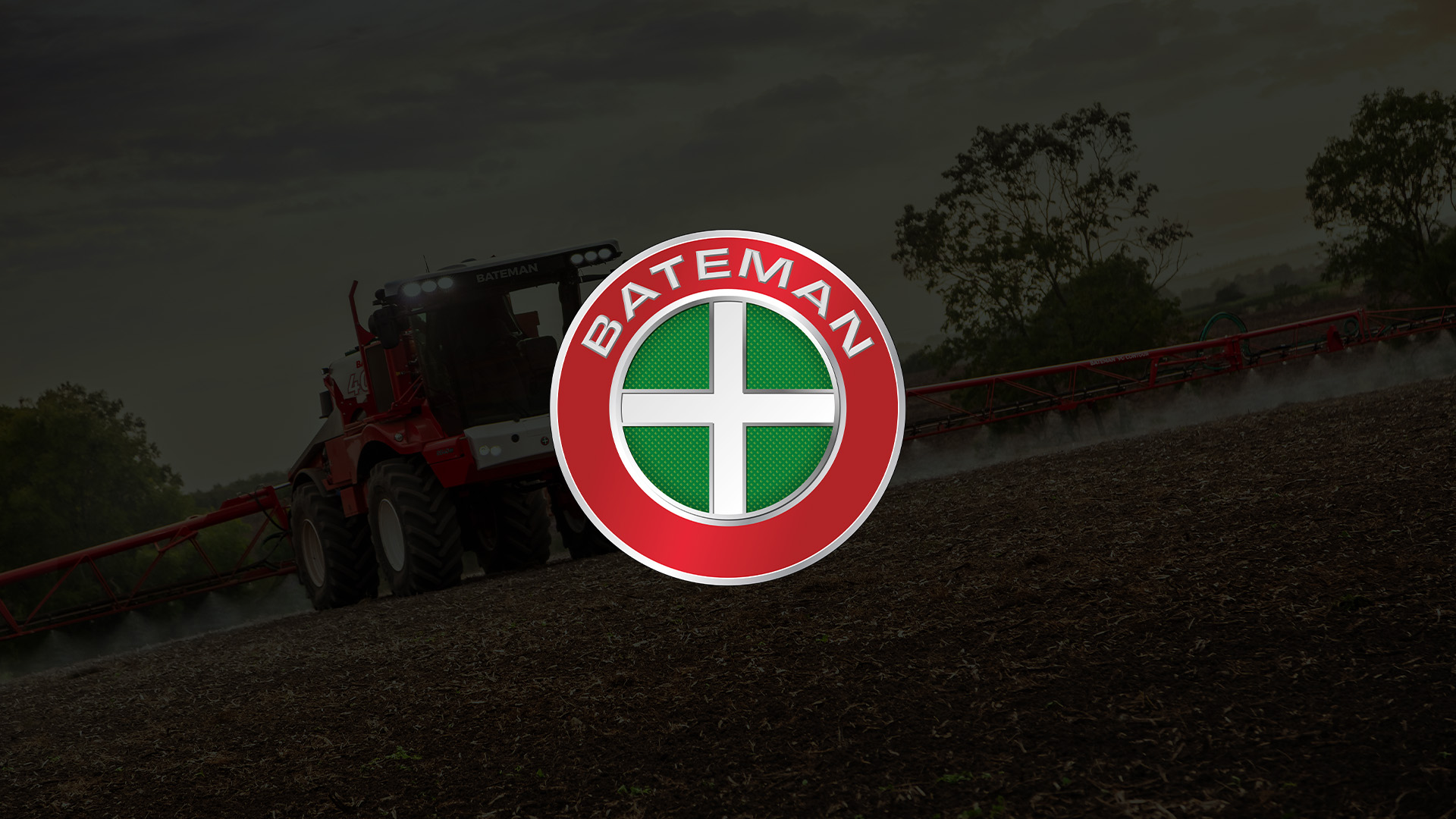 Red agricultural sprayer machine working on a field at dusk with a mist spreader arm extended, overlaid by a round Bateman logo with a green cross in the center on a red circular background.