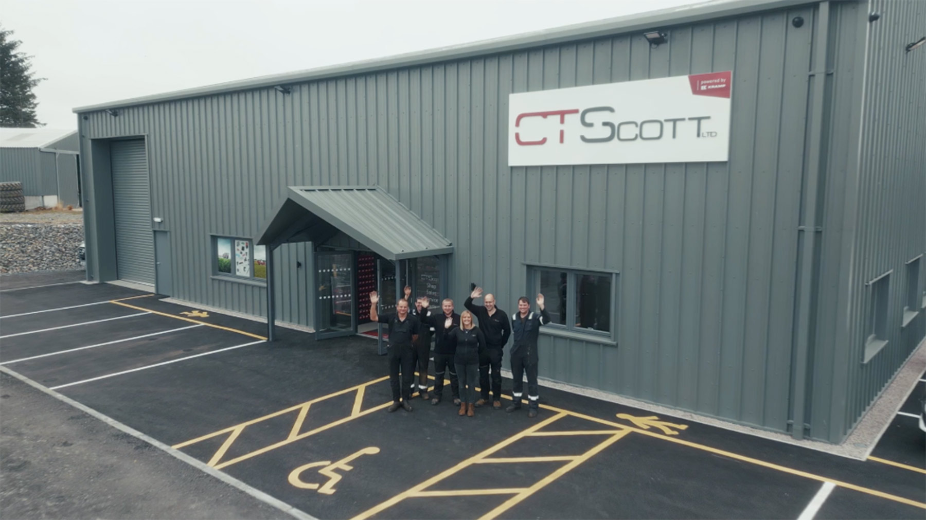 Five people standing outside the entrance of a grey industrial building labeled C T Scott Ltd, waving towards the camera, with marked parking spaces including a disabled spot in front.