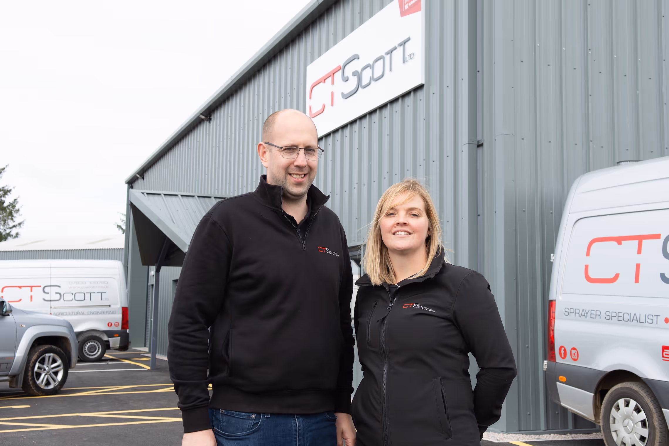 A man and a woman stand outside a gray industrial building with C T Scott signage. Both wear black jackets with the C T Scott logo. Two white company vehicles with the C T Scott logo and slogan 'Sprayer Specialist' are parked nearby.