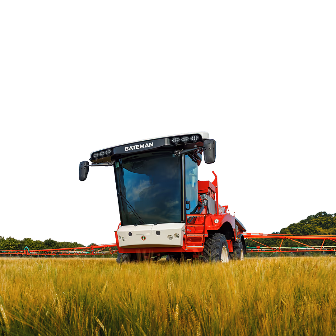 Red Bateman agricultural sprayer with large front windshield operating in a green and yellow crop field with trees in the background.