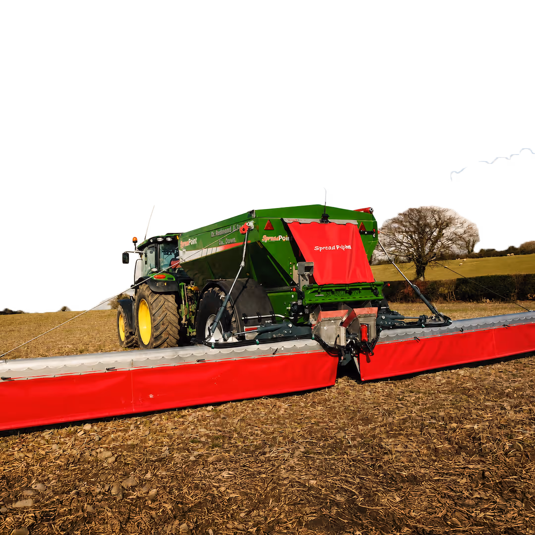 Green and red SpreadPoint agricultural spreader attached to a green tractor with yellow wheels, positioned on a plowed field with a tree and hedges in the background.