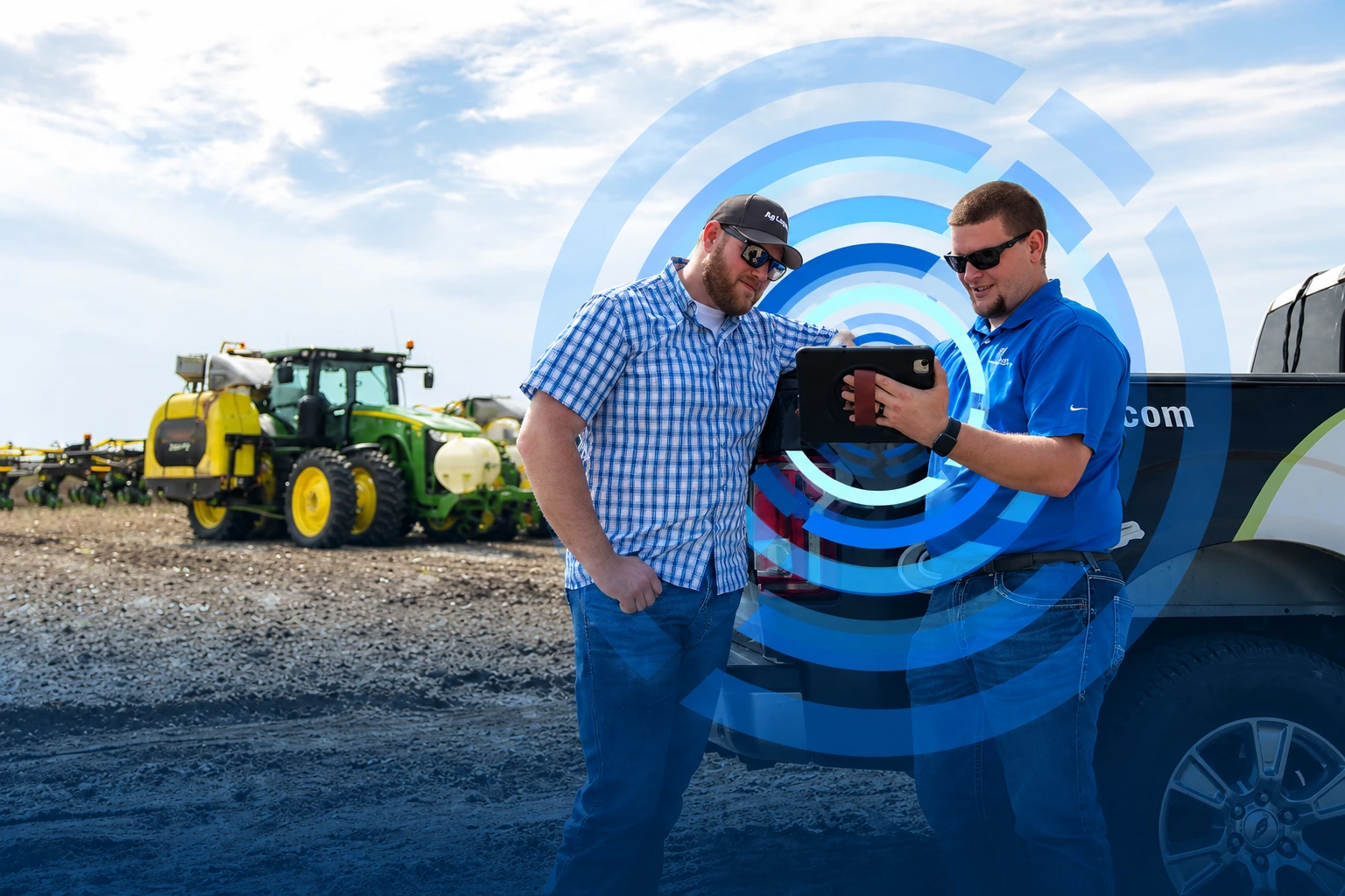 Two men wearing sunglasses and casual clothing stand next to a pickup truck in a farm field, looking at a tablet together, with agricultural machinery visible in the background.