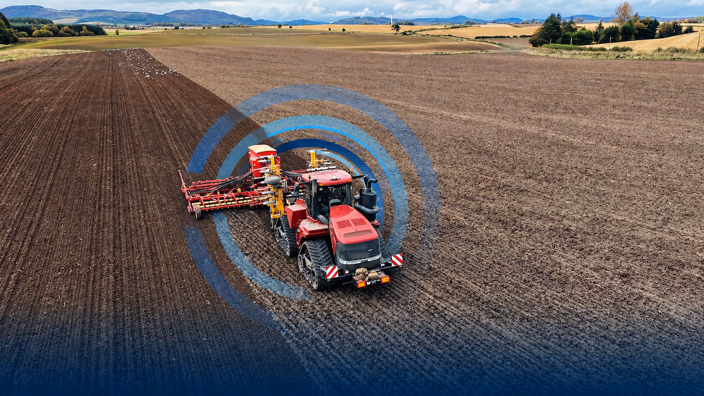 Red tractor in a large plowed field with a farming implement attached, under a partly cloudy sky with hills in the distance, overlaid with translucent blue concentric circles symbolizing connectivity or technology.