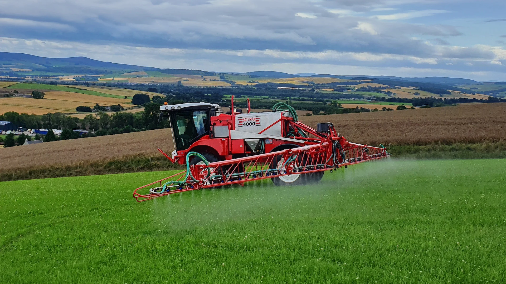 Red agricultural sprayer vehicle labeled 'Bateman 4000' applying spray over a green crop field with rolling hills and farmland in the background under a cloudy sky.