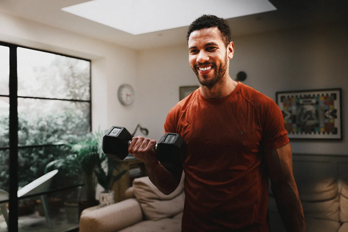 Smiling man in an orange shirt lifting a black dumbbell in a bright living room.