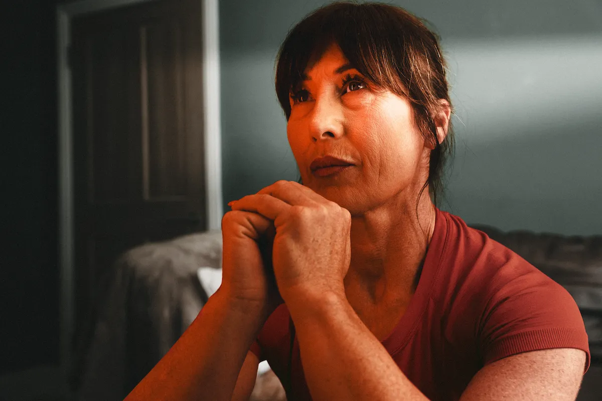 Woman with clasped hands looking upward thoughtfully, sitting indoors near a bed.