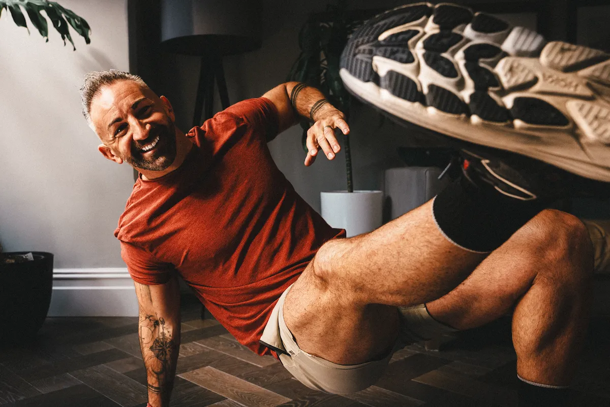 Smiling man in a red shirt exercises indoors, balancing on one arm and kicking a leg up.