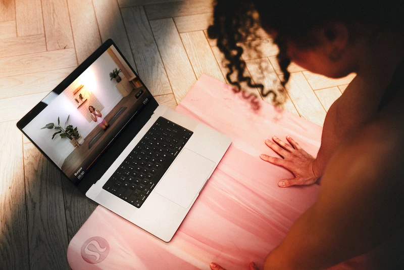 Person on pink yoga mat following an online yoga session on a laptop on wooden floor.