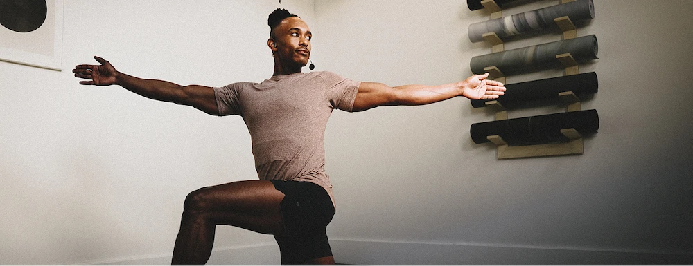 Man in athletic wear demonstrating a yoga warrior pose indoors with rolled mats on wall behind him.