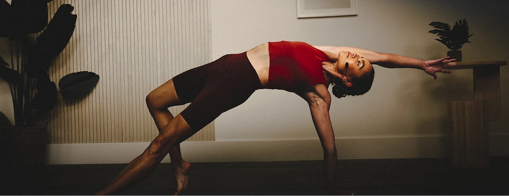 Woman in red top and shorts doing a side plank yoga pose indoors with arm extended.